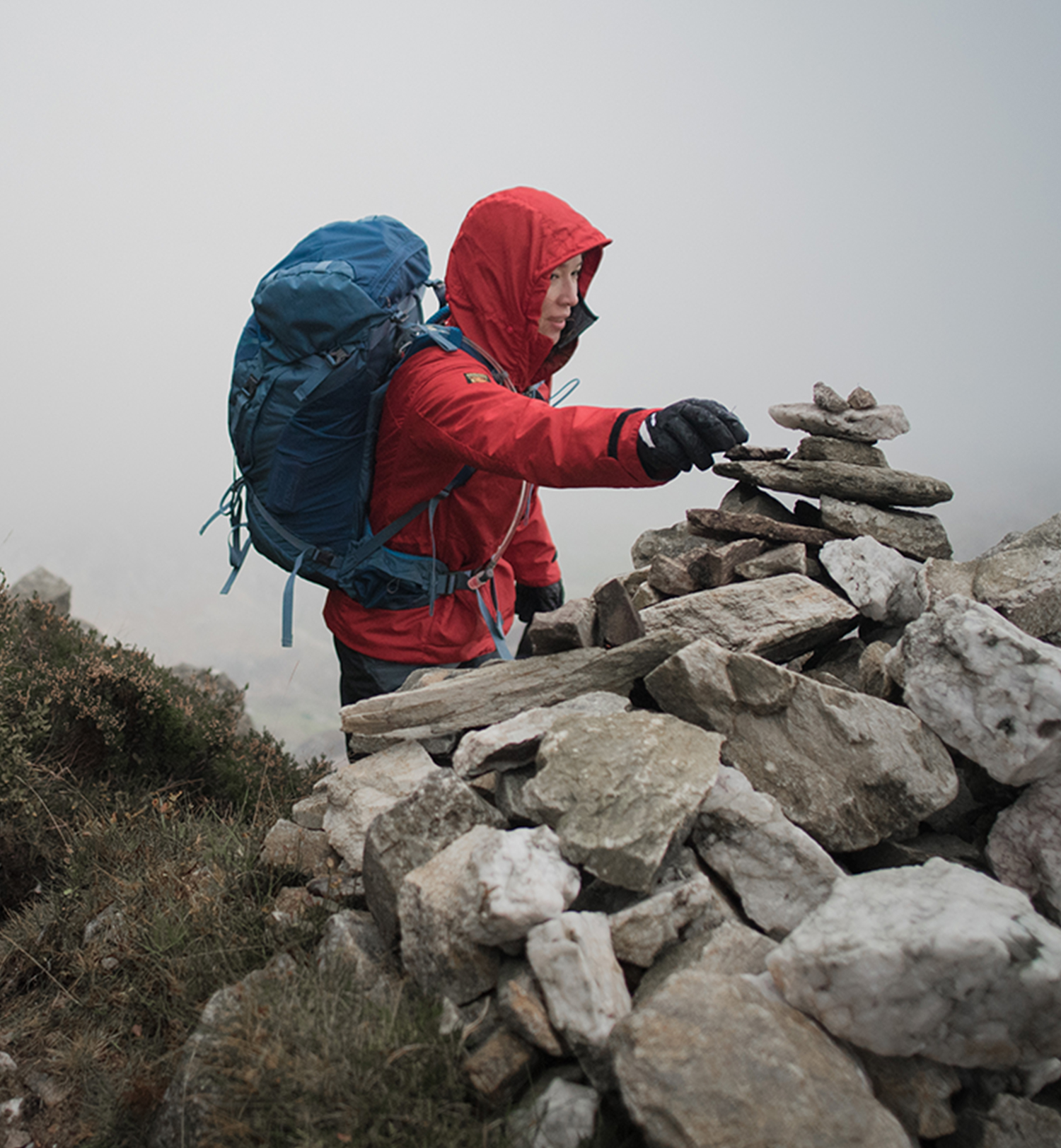 A woman in a red waterproof jacket and a rucksack places a stone on a cairn.